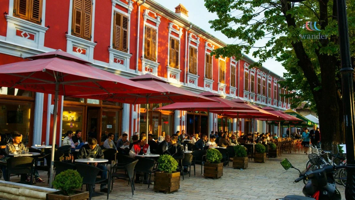 Outdoor dining area on a city street with tables, chairs, and large umbrellas, in front of a colorful red and white historic building, with people seated enjoying the evening.