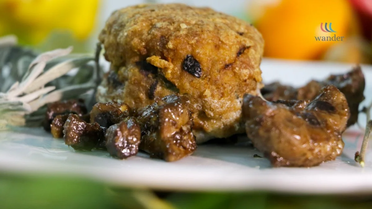 Close-up of a fried chicken leg with a crispy coating, served with cooked, glazed mushrooms on a white plate. Blurred colorful background.