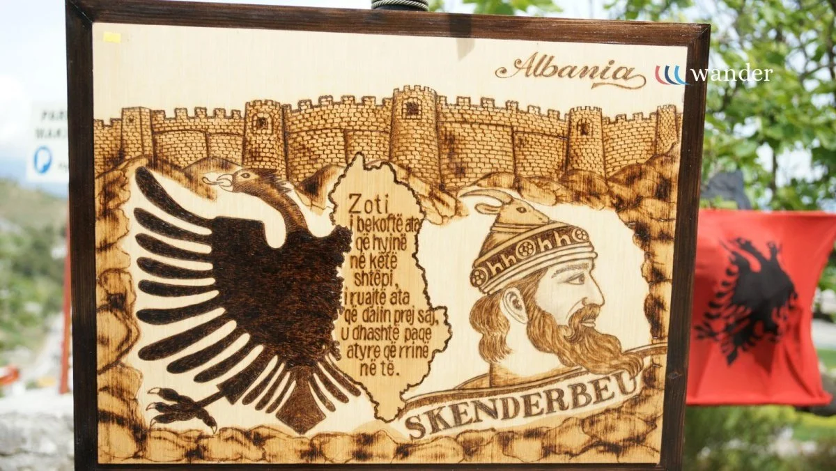 Wooden plaque featuring an illustration of a castle, an eagle, and a historical figure with a crown and beard. The plaque has text in Albanian referencing Skenderbeu, a national hero, with the word "Albania" and the logo "wander" in the top right corner.
