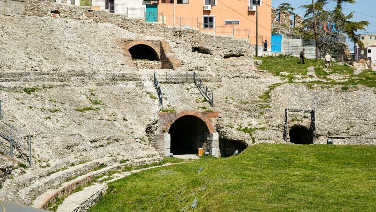 Historical ruins with arched entrances built into a hillside, with a grassy foreground and buildings in the background.