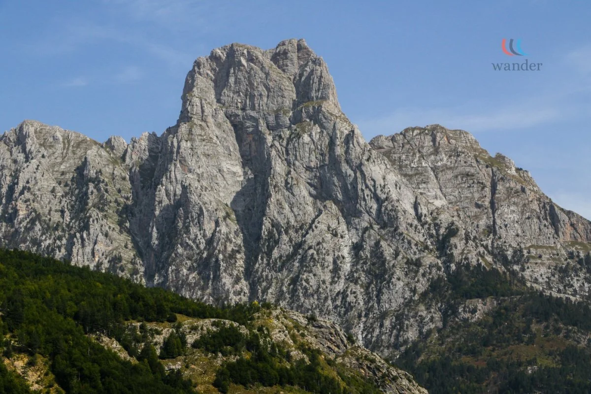 A large rocky mountain with steep, rugged cliffs and a forested base, under a blue sky with some wispy clouds.
