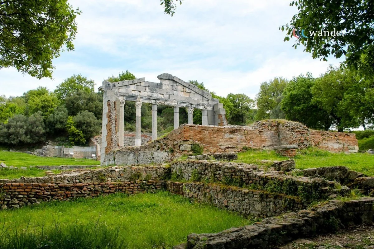 Ancient Greco-Roman ruins with columns and brick walls surrounded by green trees and grass, under a light blue sky.