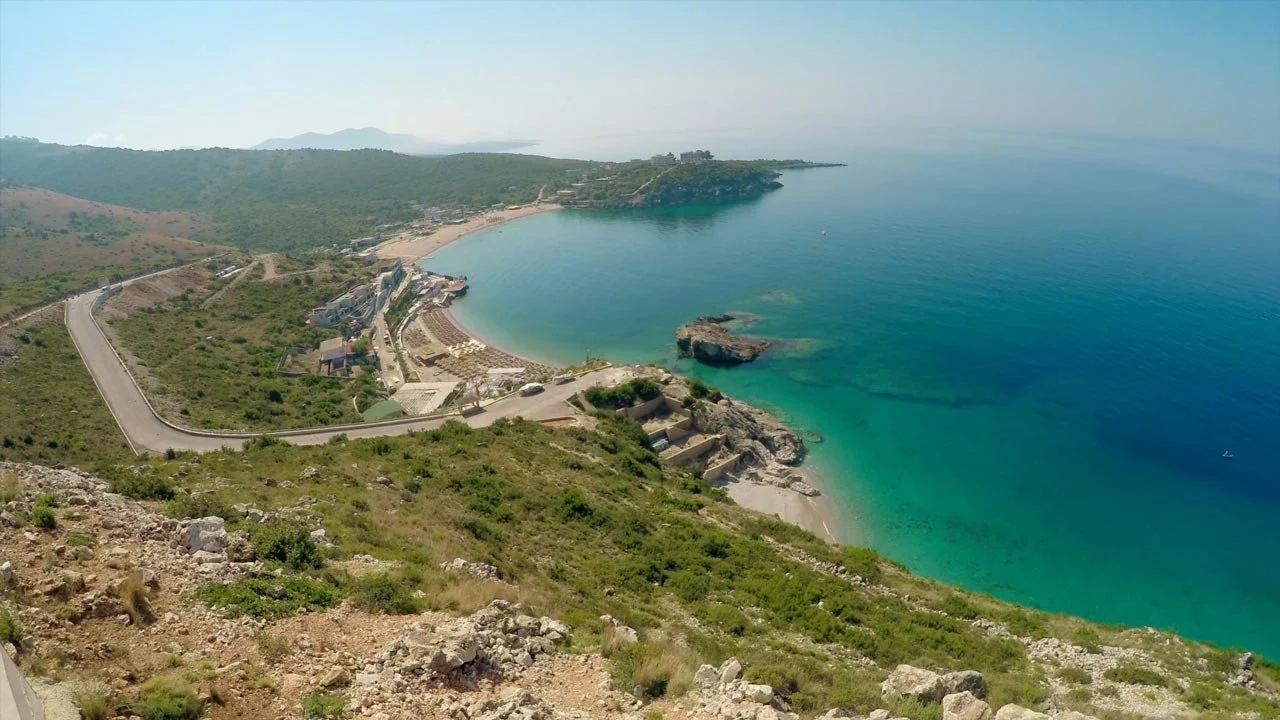 Aerial view of a crescent-shaped beach with turquoise water, rocky outcroppings, and a coastal road lined with buildings and greenery.