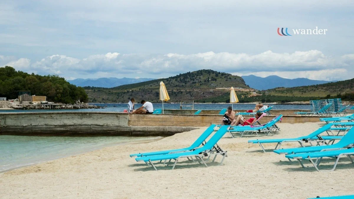 Beach scene with blue lounge chairs on sand, a few people relaxing by the water, and a dock with umbrellas in the background. Mountains and partly cloudy sky visible in the distance.
