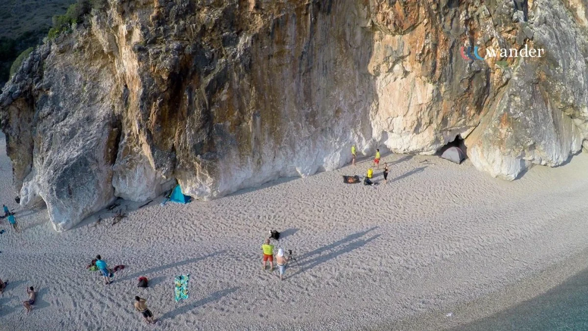 Beach scene with people, tents, and camera setup near large rocky cliffs.
