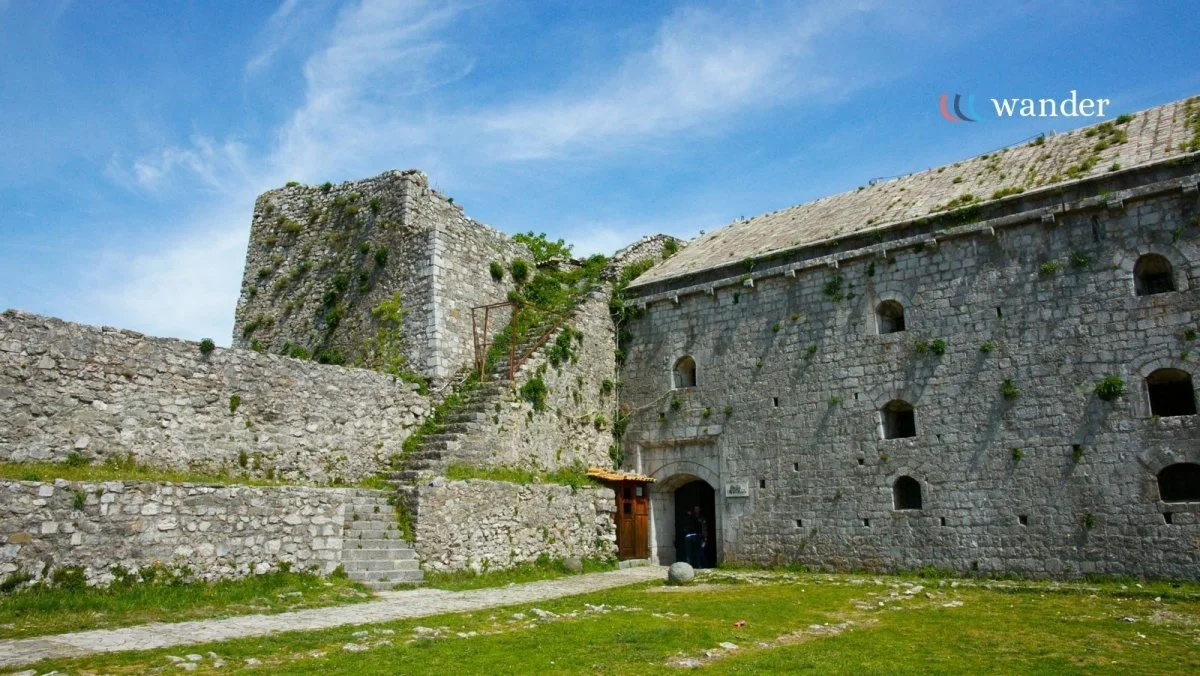 Old stone castle with stairs and lush grass in front, blue sky with clouds above.