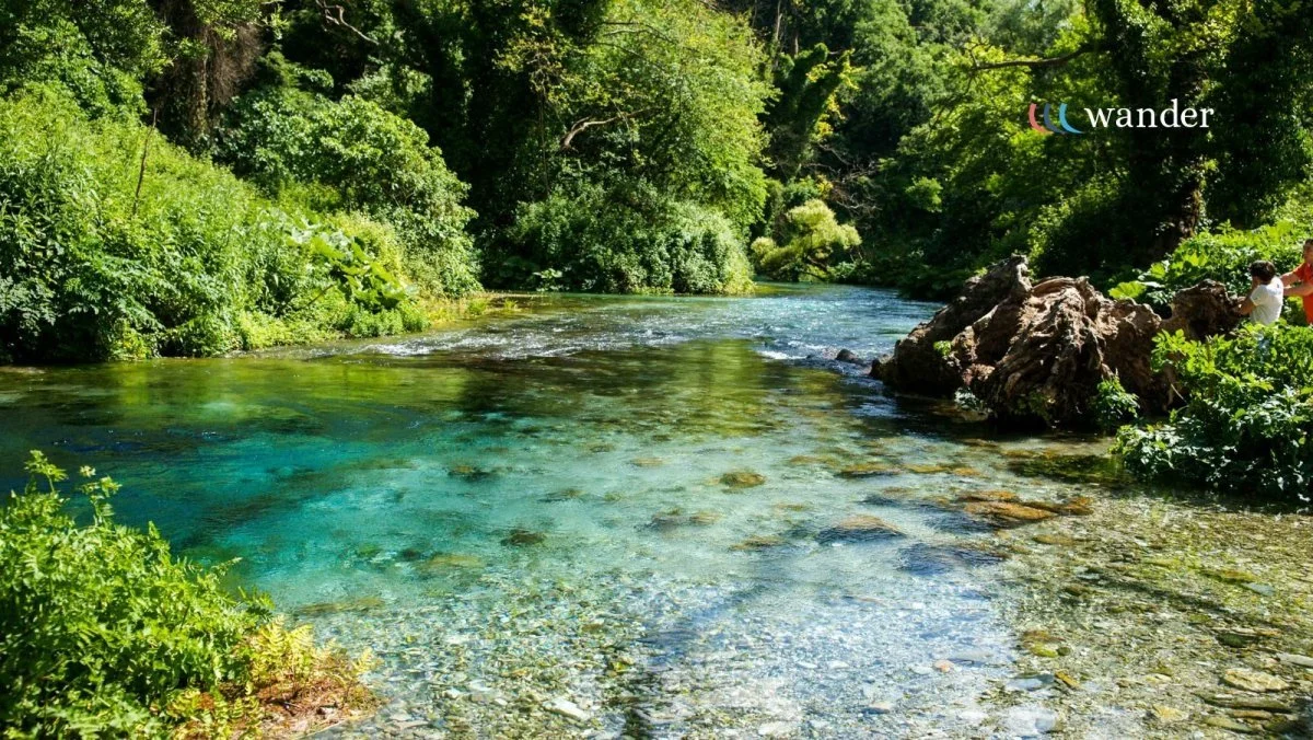 A clear, turquoise river flowing through a lush, green forest with dense trees and bushes on both sides. There are two children on the right side near the rocks.