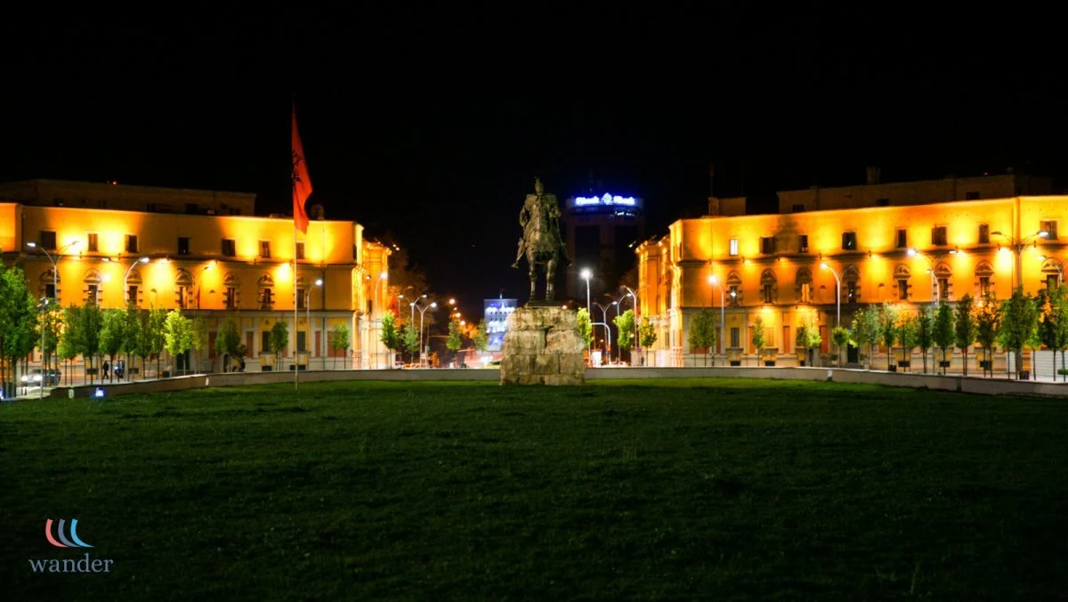 Nighttime view of a city square with a central statue of a man on a horse on a pedestal, surrounded by illuminated buildings and trees, with a flag on a pole to the left.