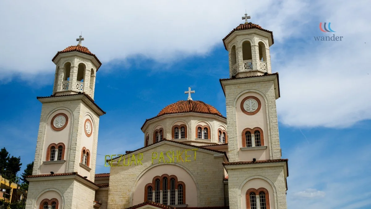 A church with three towers topped with crosses, a central dome, and a yellow sign that reads "SEZUAR PASHAK" against a blue sky with clouds.