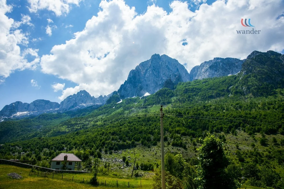Green mountains with rocky peaks under a partly cloudy sky, a house, and a power line in the foreground.