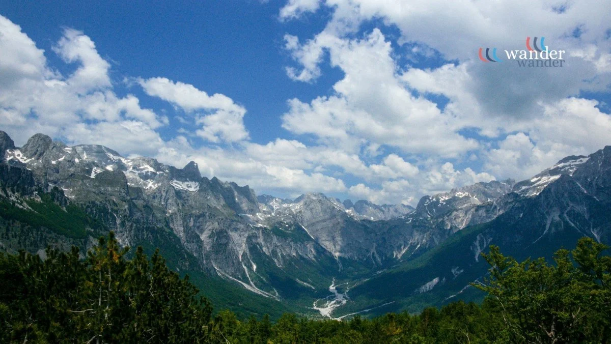 Mountain landscape with snow-capped peaks, green valleys, and a partly cloudy sky.