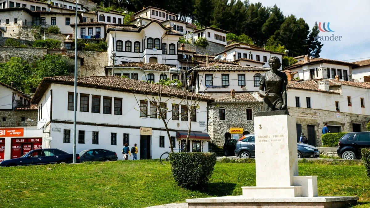 A statue of a woman stands on a pedestal in a park with grass and bushes. In the background, there are white buildings with tiled roofs on a hillside, cars parked along the street, and a few people walking.