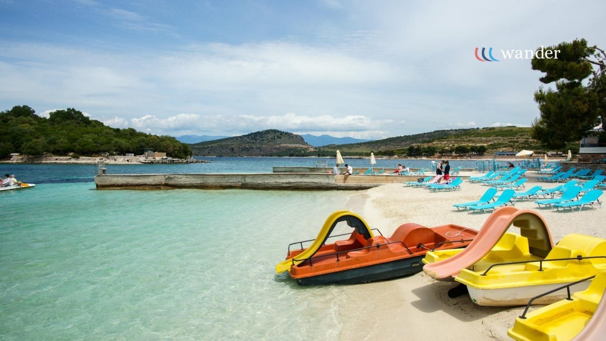 Sunny beach with colorful paddle boats on the shore, rows of blue lounge chairs, and a few people relaxing near the water with hills and a cloudy sky in the background.