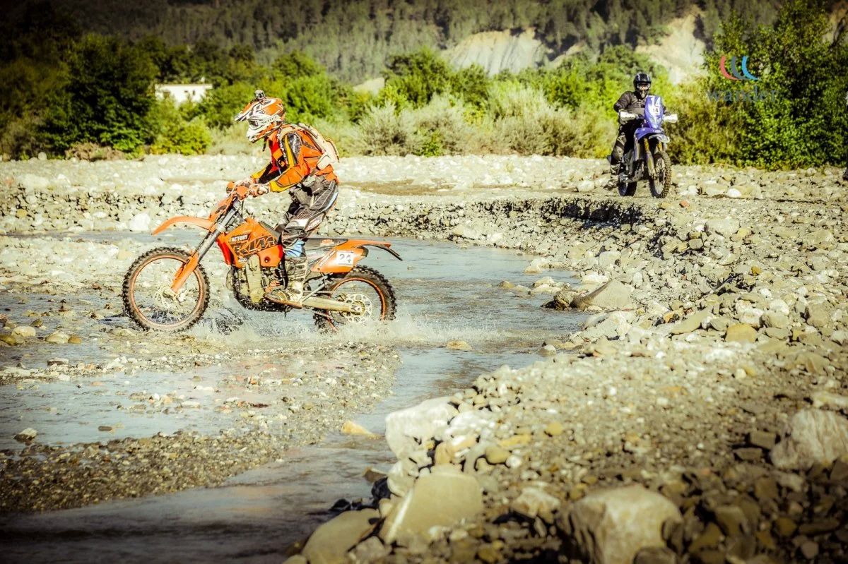 Two motorcyclists crossing a shallow river on a rocky terrain in a rural area with green trees and distant hills.