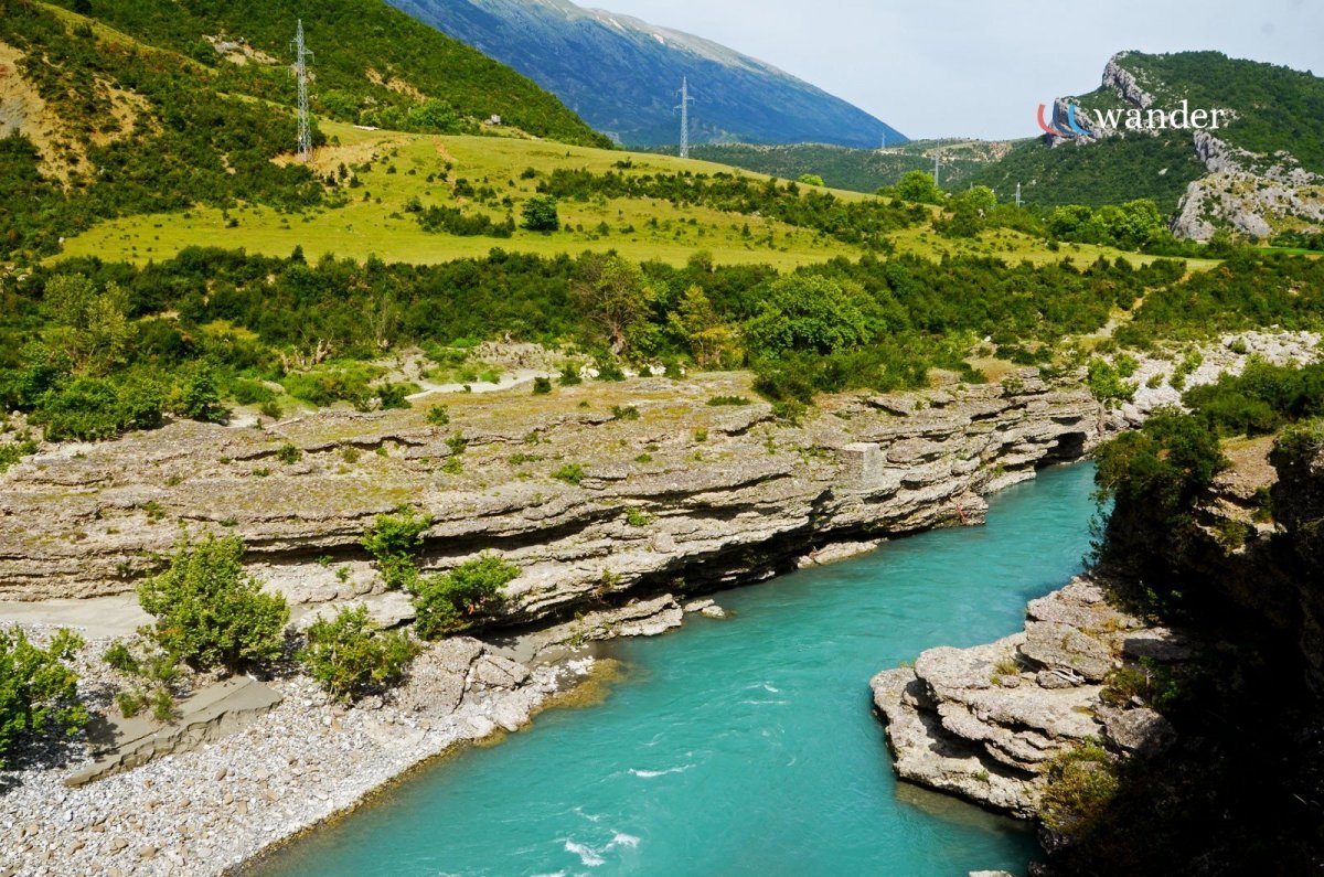 A scenic view of a turquoise river flowing through a rocky canyon surrounded by lush green hills with some power lines and mountains in the background.