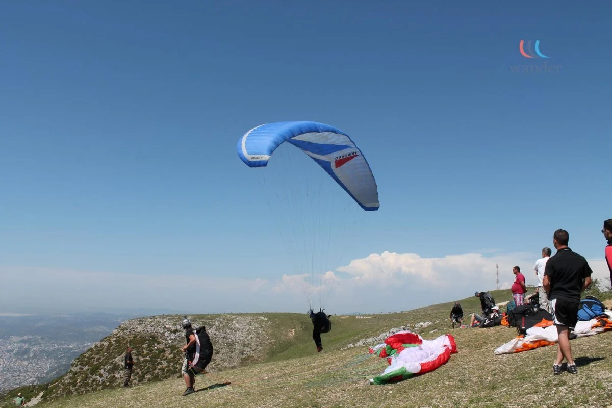 People on a grassy hill preparing for paragliding, with one person taking off with a blue and white parachute, and others watching or sitting near their gear.