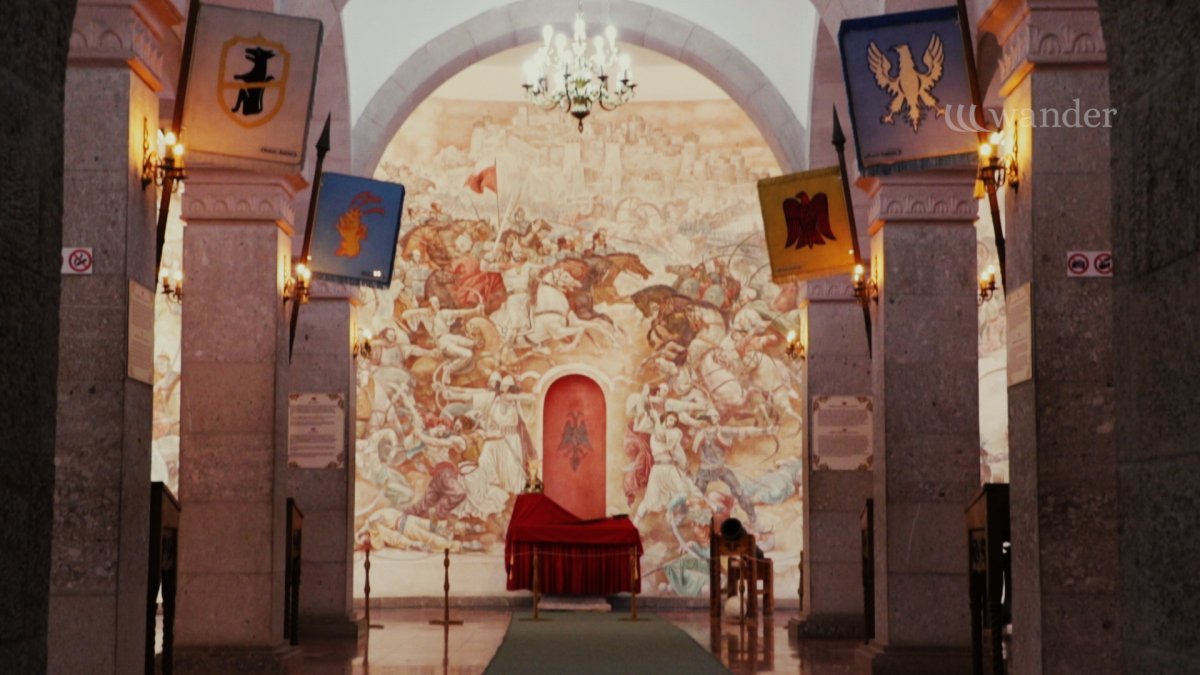 Interior of a historic building with murals depicting a battle scene, medieval banners hanging from columns, and an altar covered with a red cloth at the center.