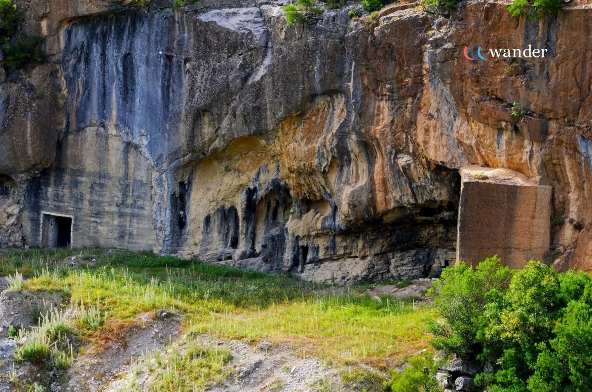 Rocky cliffside with caves and green vegetation at the base, logo with the word 'wander' in the top right corner.