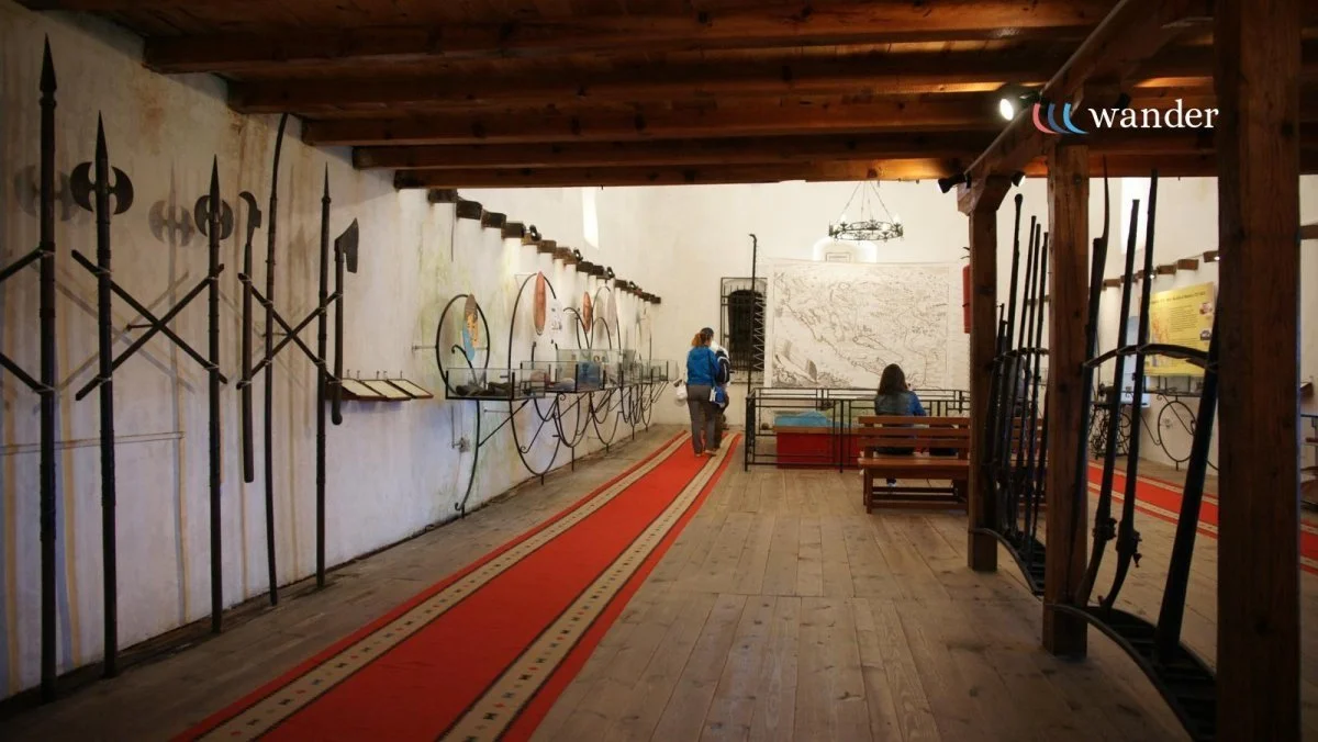Interior of a historical museum with display swords and spears on the left wall, a red carpet running through the center, and visitors viewing exhibits.