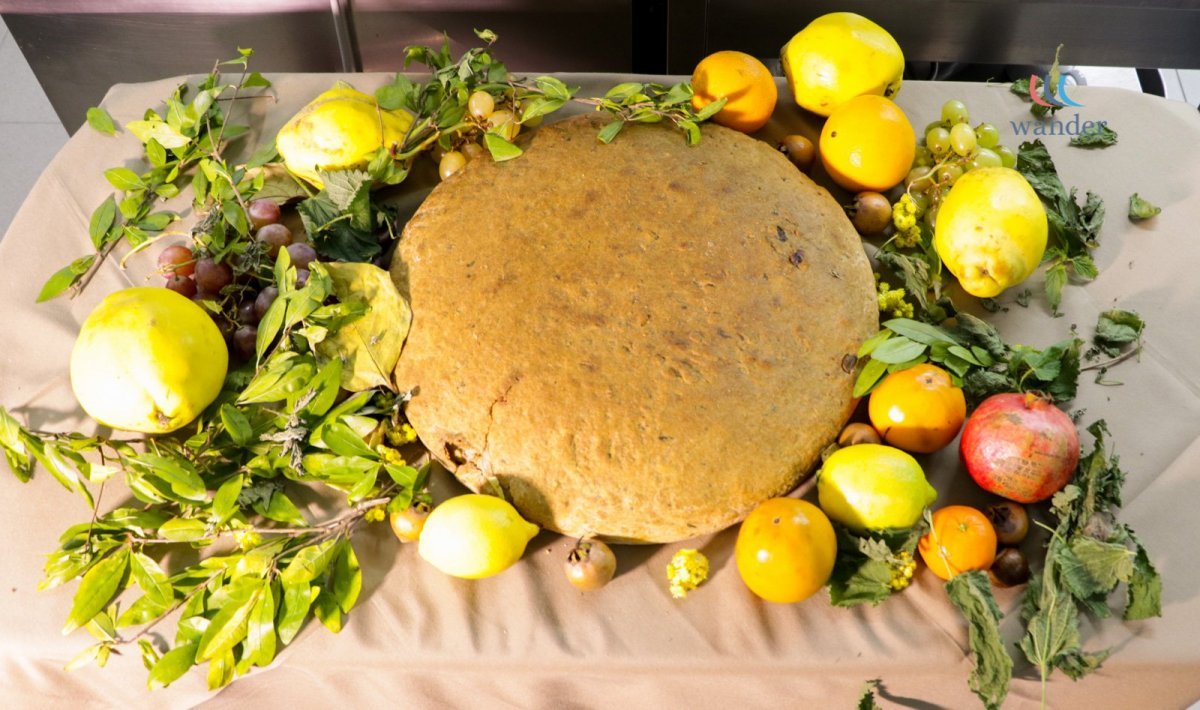 A large round loaf of bread surrounded by various fruits and green foliage on a table.