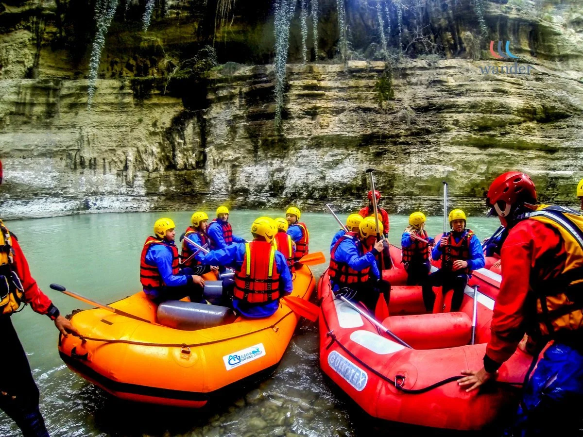 Group of people in yellow helmets on orange and red inflatable rafts preparing for whitewater rafting in a river surrounded by tall cliffs.