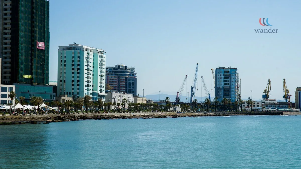 A waterfront cityscape with tall modern buildings, construction cranes, palm trees, and a promenade along the water.