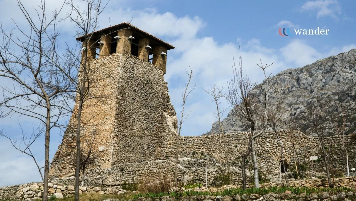 Ancient stone tower with a wooden roof, surrounded by leafless trees and a rocky mountain in the background, under a partly cloudy sky.