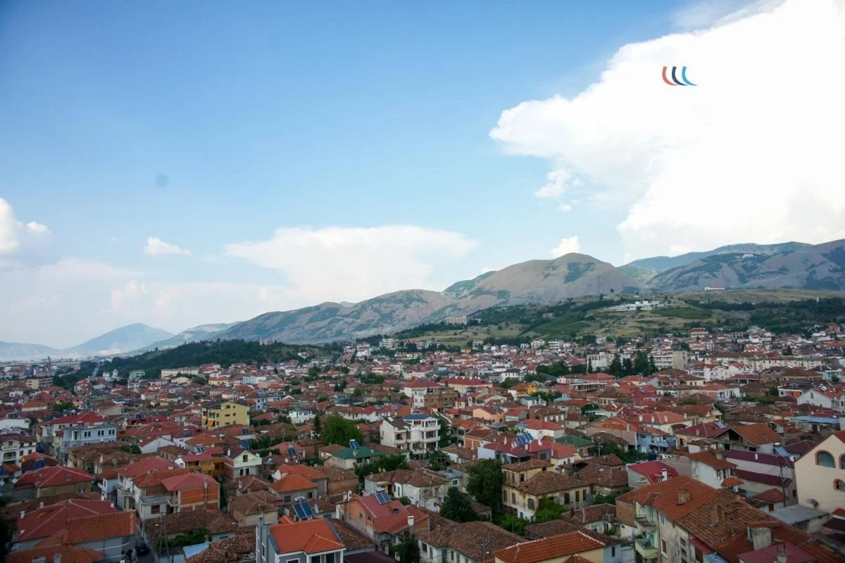 A panoramic view of a city with red-tiled roofs, surrounded by green hills and mountains under a partly cloudy blue sky.