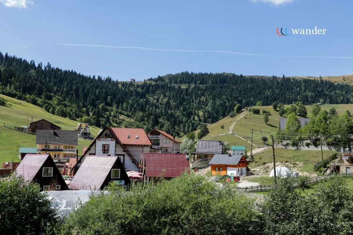 A scenic view of a small hillside village with colorful houses surrounded by green trees and rolling hills under a blue sky.