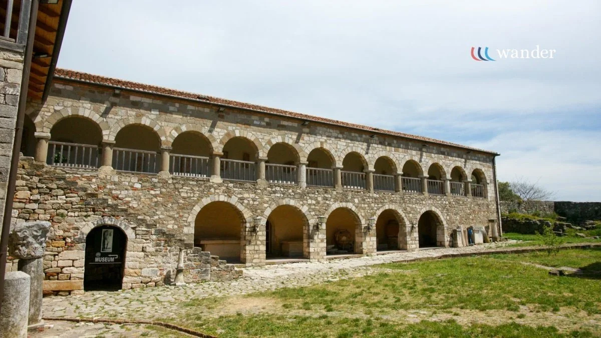 Ancient stone building with arches and two floors, with a museum entrance on the ground level, surrounded by grass and an overcast sky.