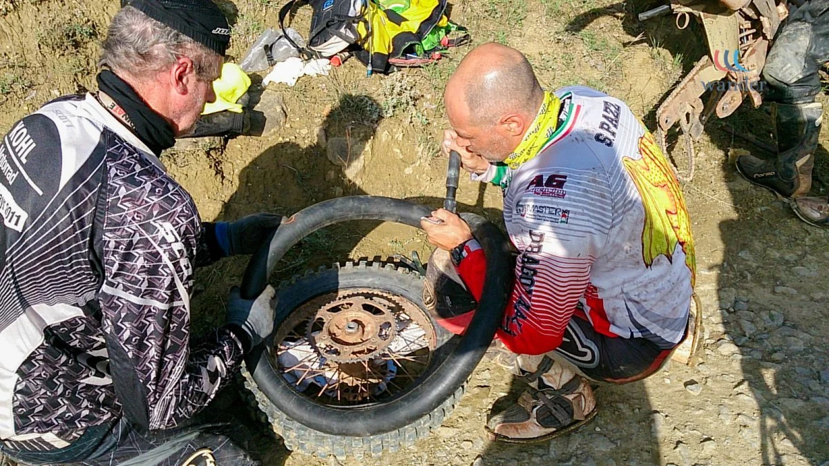 Two men dressed in motocross gear work on a motorcycle tire on a dirt surface. One man is holding the tire, and the other is inspecting or adjusting the wheel. There are motocross backpacks and gear in the background.