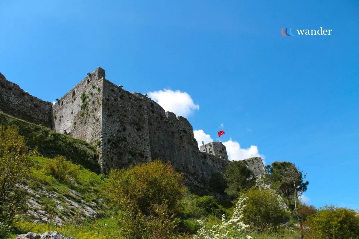 A historic stone fortress with a flag on top, situated on a hillside with green trees and shrubs, under a blue sky with some clouds.