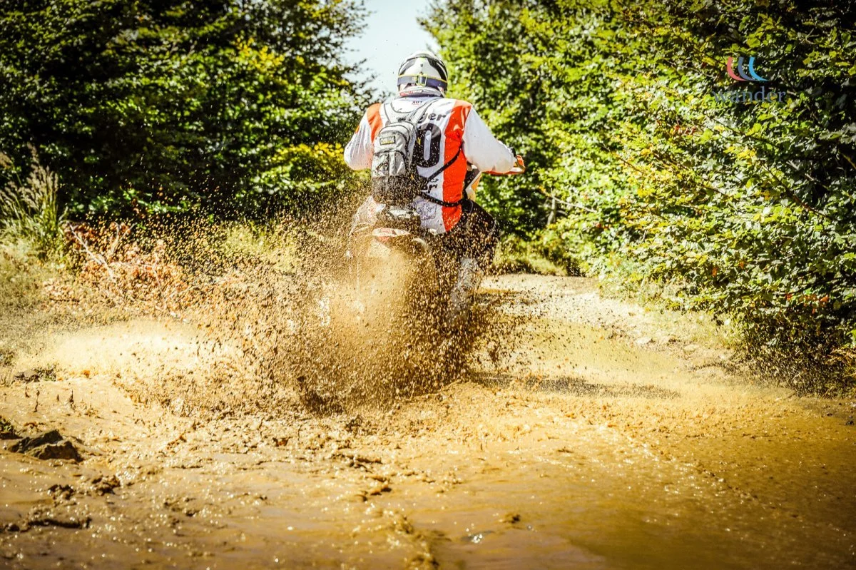 A person riding a dirt bike through a muddy trail surrounded by green trees.