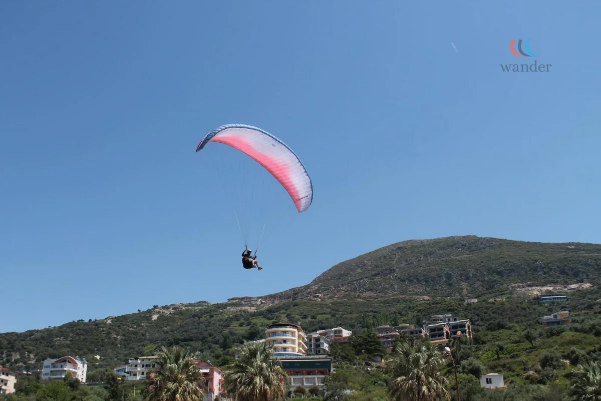 A person paragliding with a pink and white parachute over a hillside with houses and palm trees under a clear blue sky.