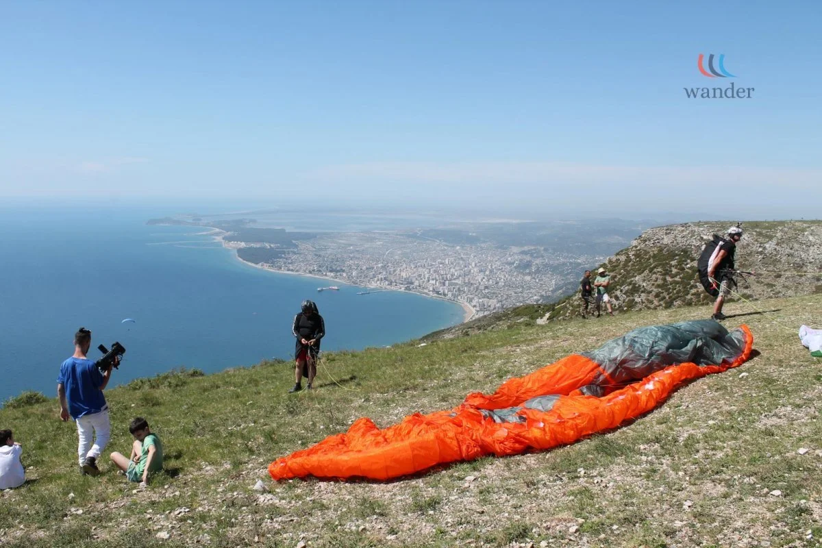People preparing for paragliding on a grassy hillside with a view of the ocean and city in the background, some children sitting on the grass.