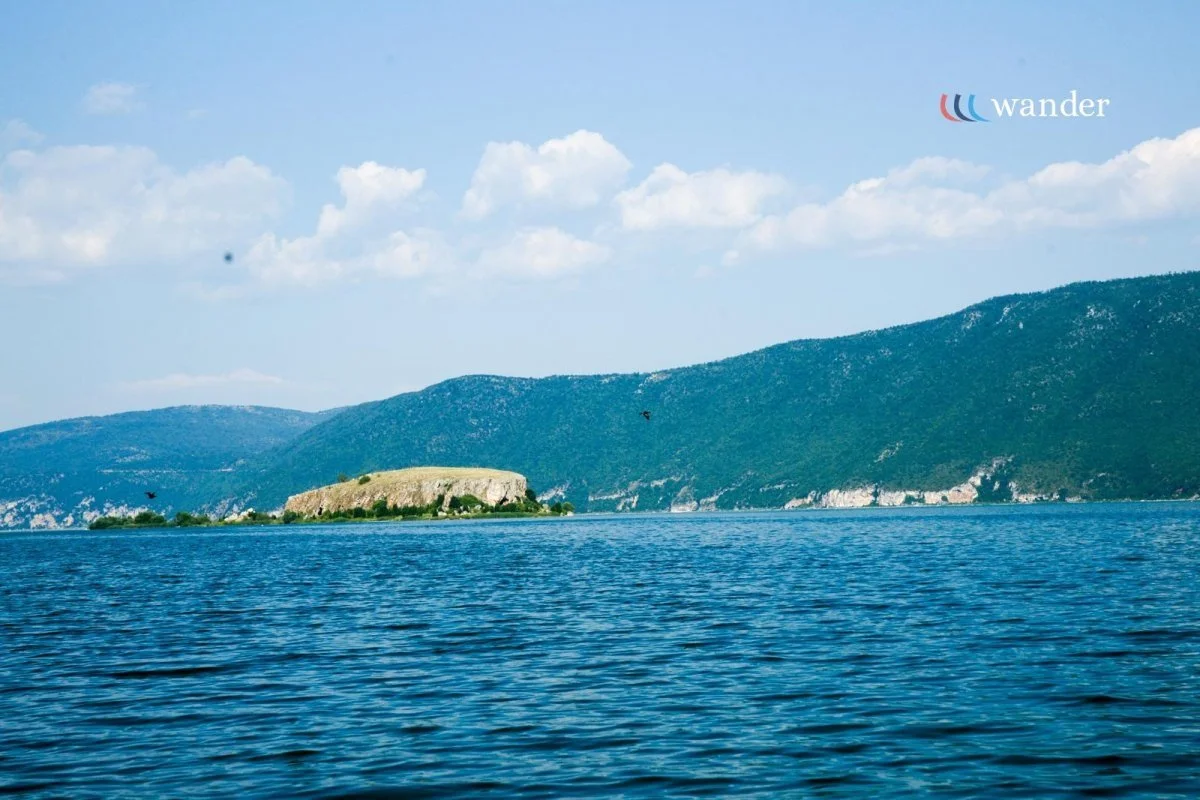 A scenic view of a large body of water with distant hills and a small island in the middle. The sky is partly cloudy with white fluffy clouds. The word 'Wander' is in the top right corner.