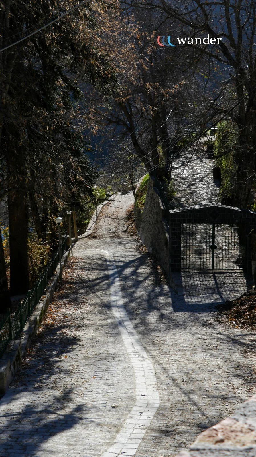 A winding cobblestone pathway lined with trees and a black metal gate in a shaded, wooded area.