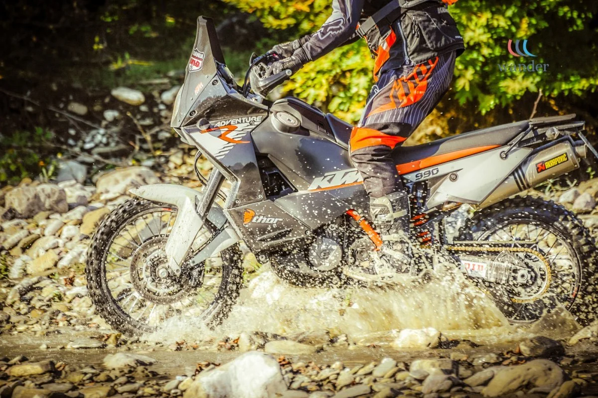 A person riding a black and orange KTM Adventure motorcycle through a rocky stream, splashing water as they ride.