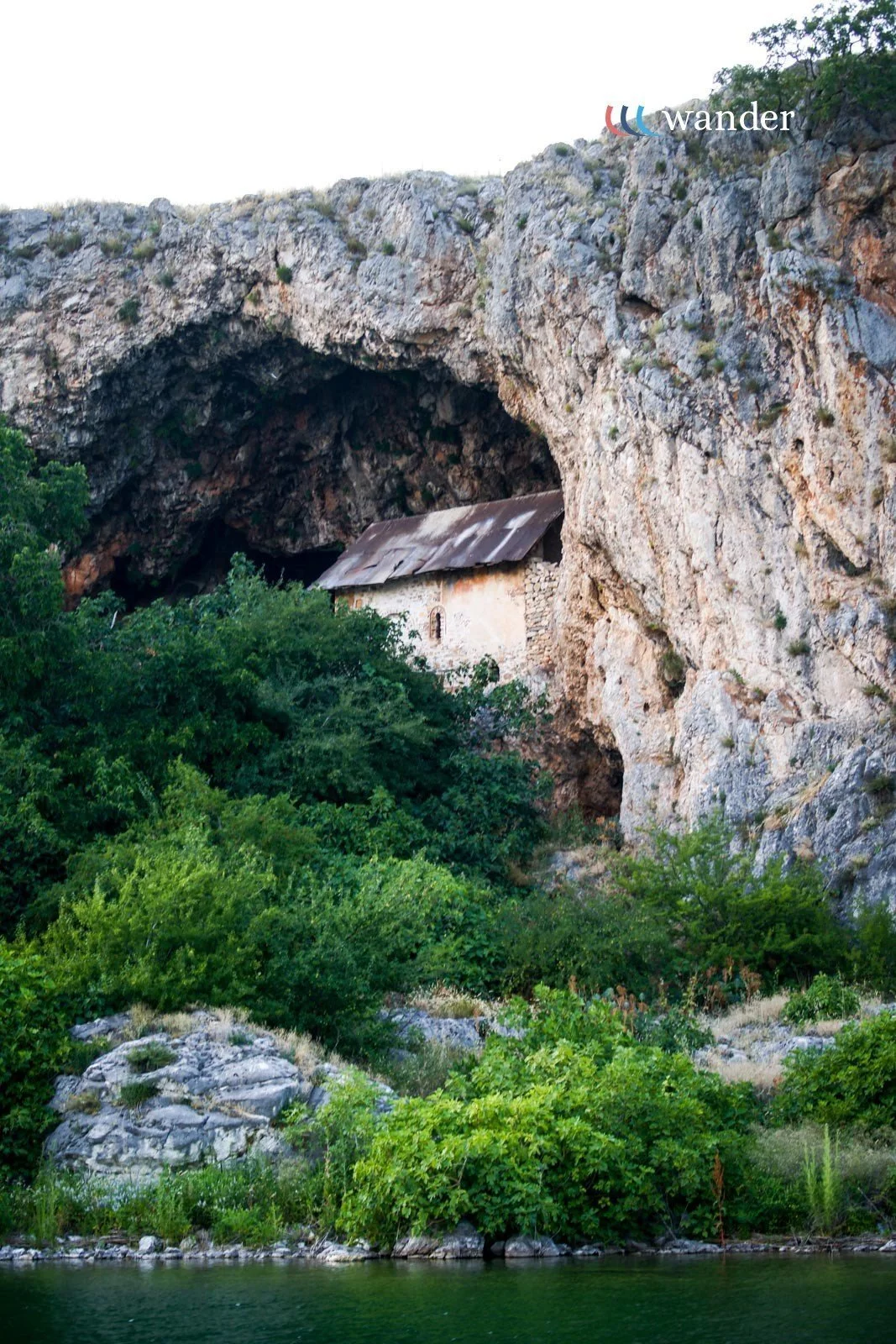 A small house with a corrugated metal roof nestled in a rocky hillside surrounded by lush green bushes and trees, with a body of water in the foreground.
