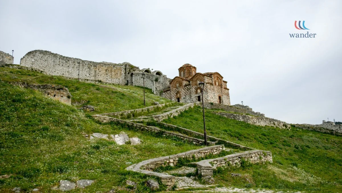 Ancient stone fortress on a grassy hillside with a zigzag stone pathway leading to a small church at the top. Overcast sky.
