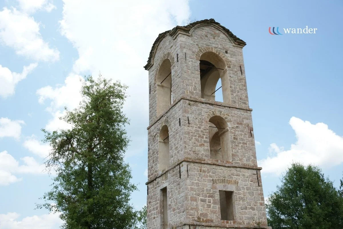 Stone tower with three arched openings on each of its four levels, surrounded by green trees and a partly cloudy sky.