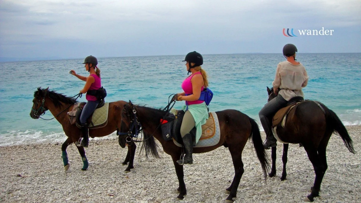 Three women riding horses along a pebble beach near the ocean, with the waves and sky in the background.