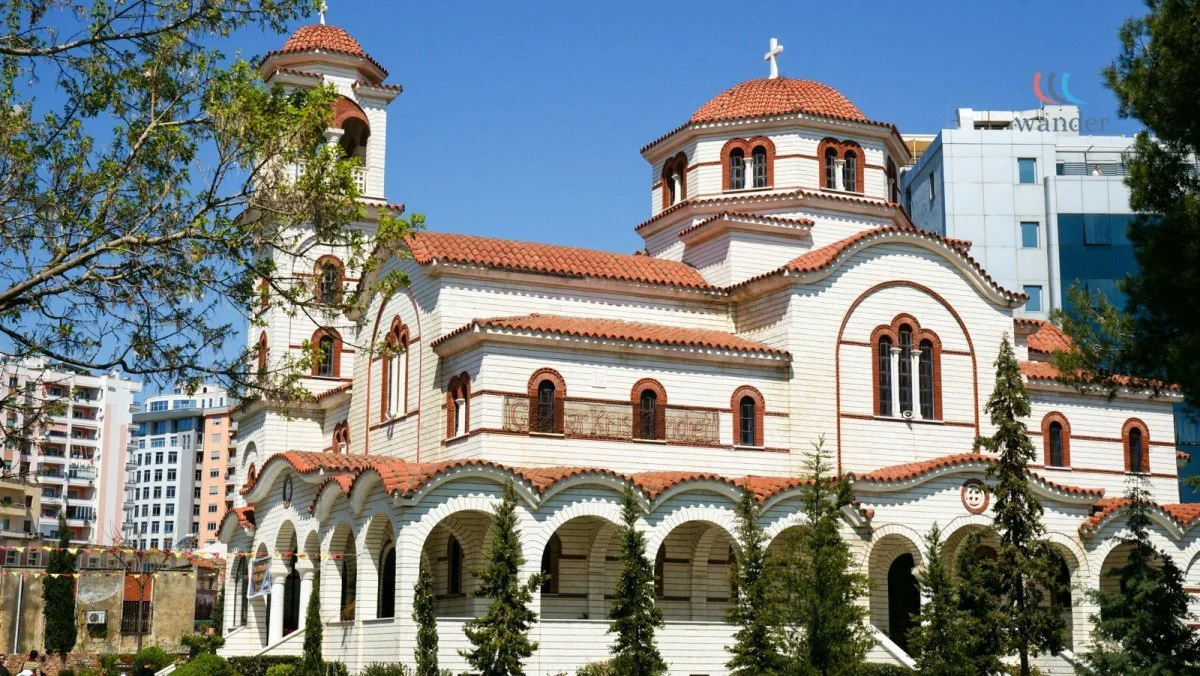 Orthodox church with red tiled domes and white walls, surrounded by trees, in an urban setting with modern buildings in the background.