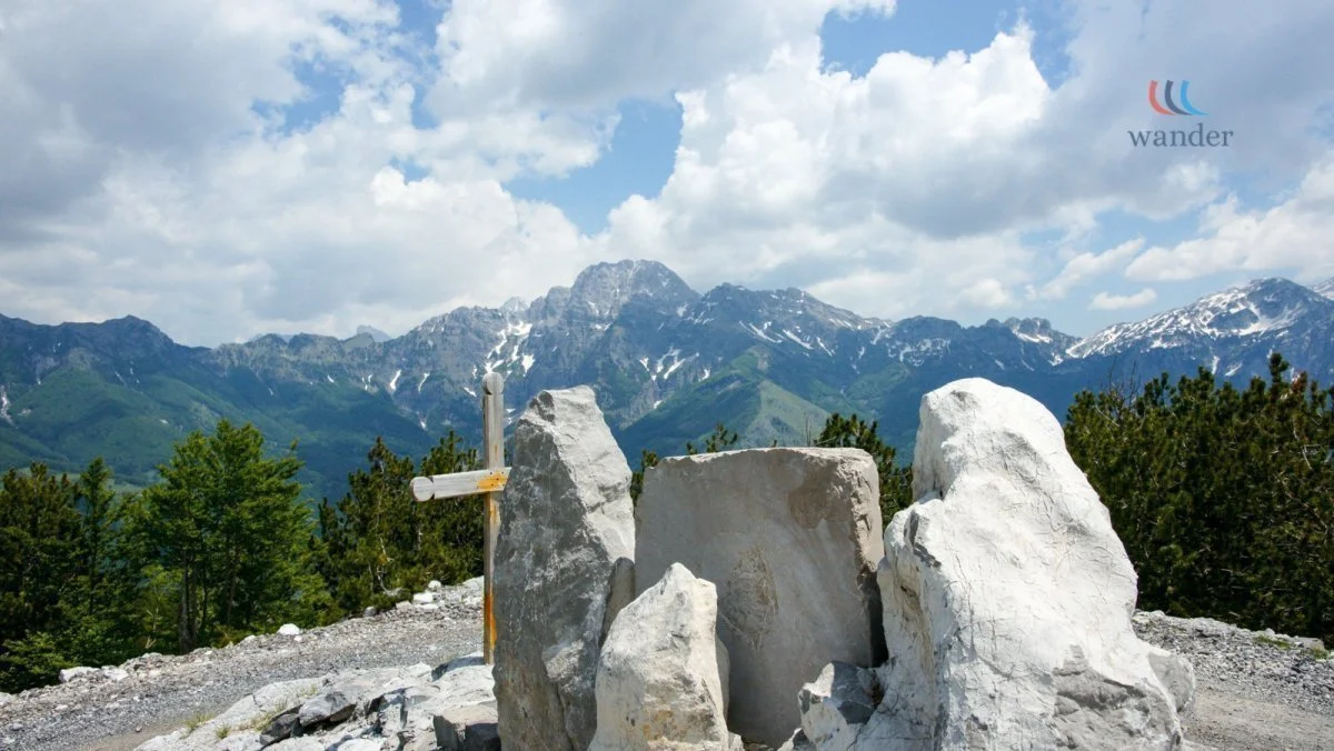 A large rock formation in a mountainous landscape with green trees, snow-capped peaks, and a partly cloudy sky in the background.