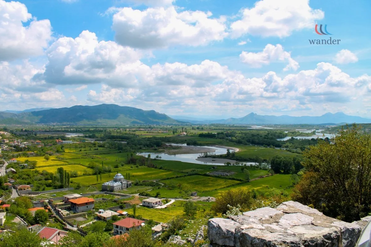 Scenic view of a lush green valley with rivers, houses, and mountains under a partly cloudy sky.