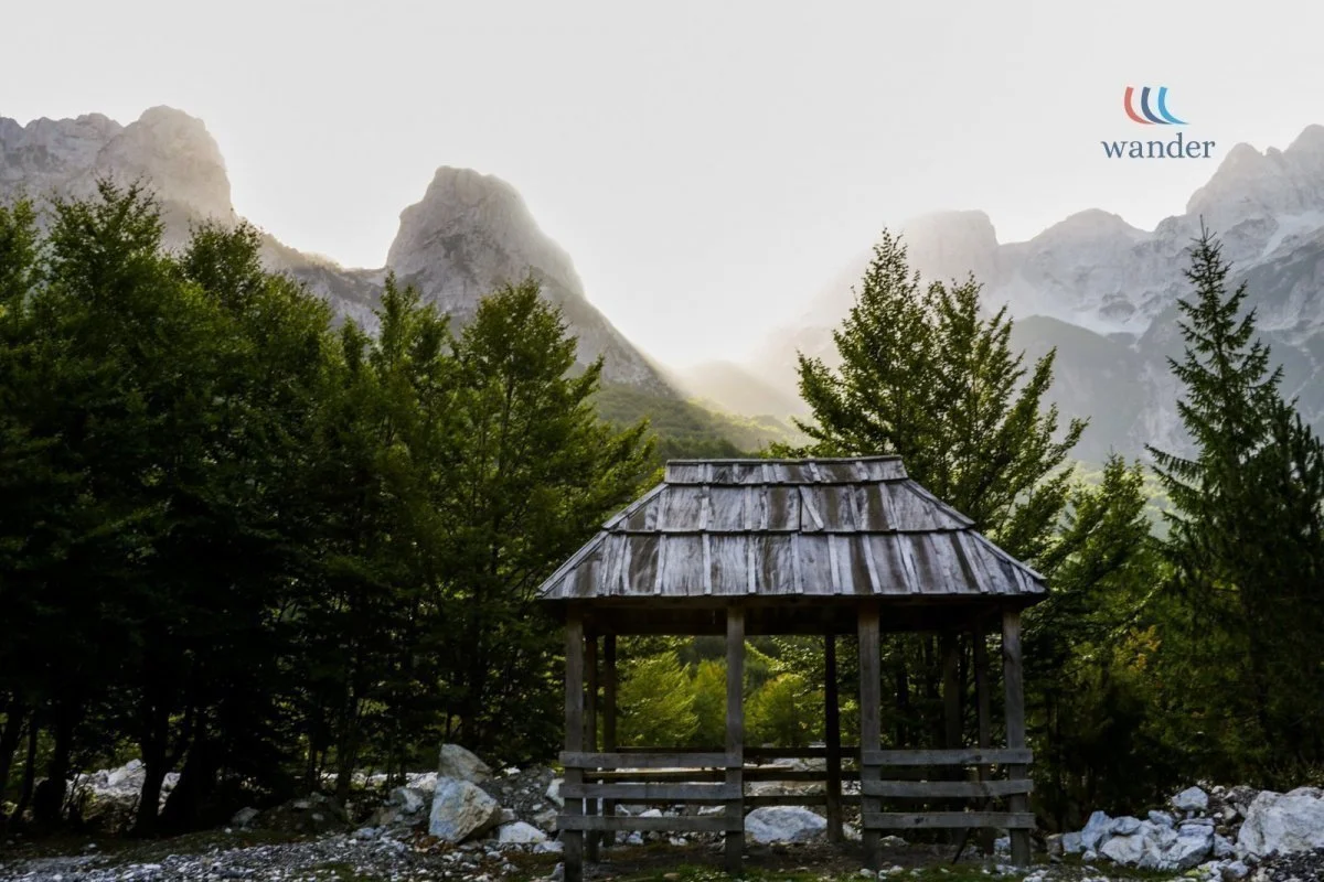 A wooden shelter with a weathered roof in front of a dense forest with tall trees, and mountains in the background with snow on their peaks.