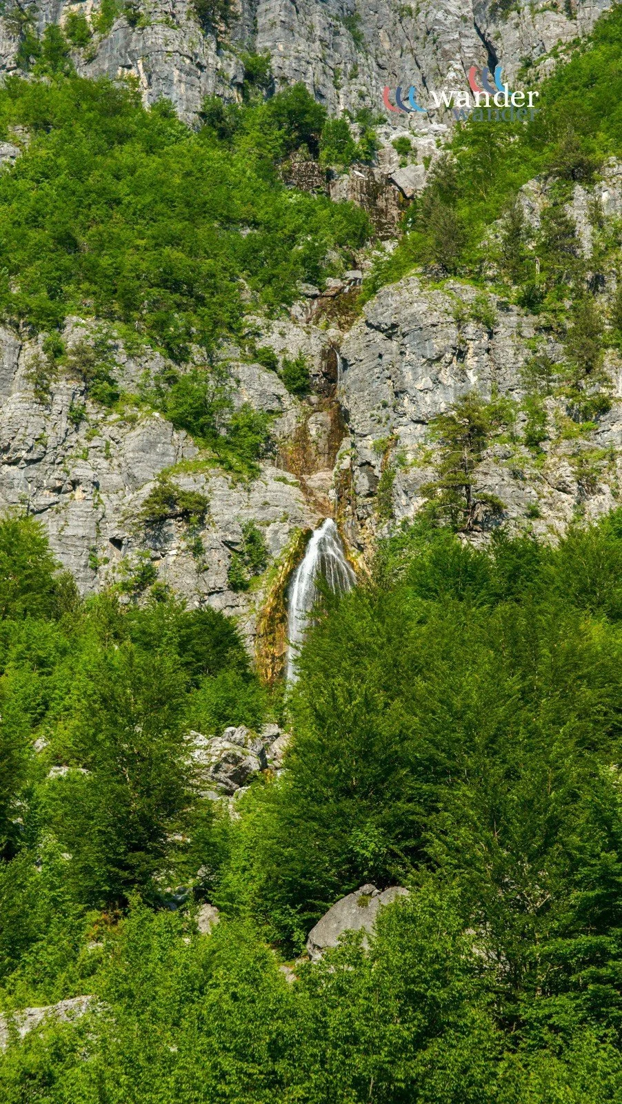 A scenic view of a narrow waterfall cascading down a rocky cliff surrounded by lush green trees.