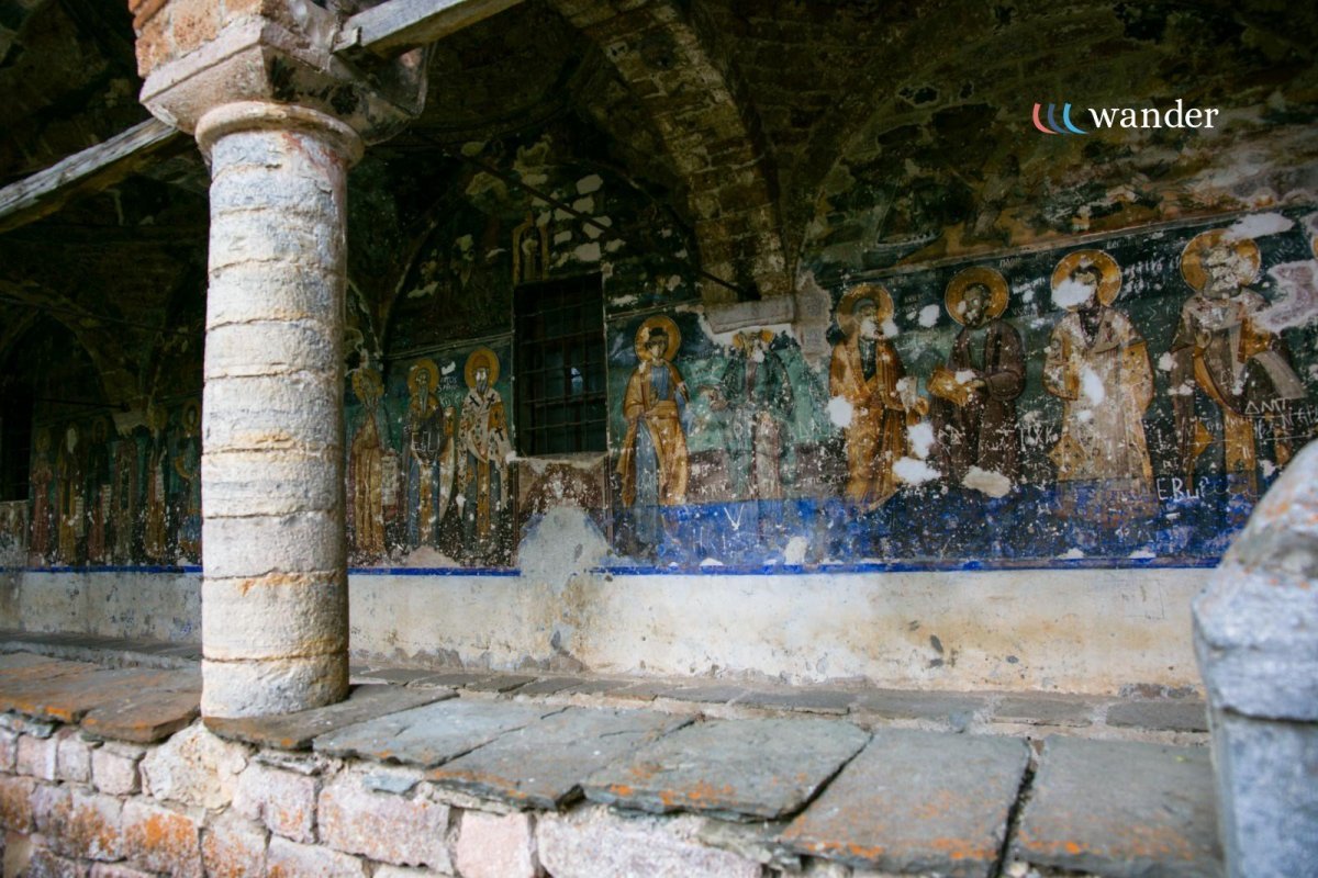 Ancient fresco of saints and religious figures on a weathered wall inside a stone structure with a brick floor and column. The fresco's paint is chipped and faded.