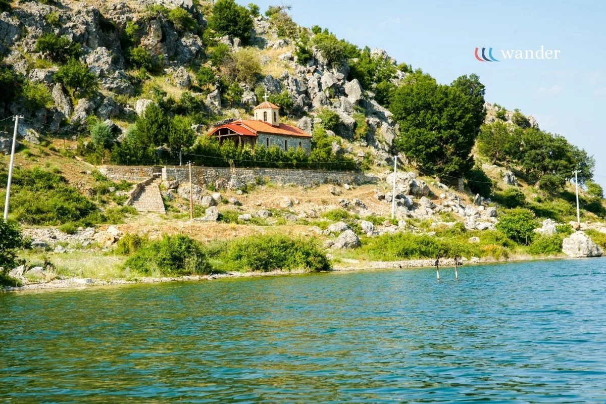 A lakeside scene with a rocky hillside, green trees, a small building with a red roof, and power lines near the water.