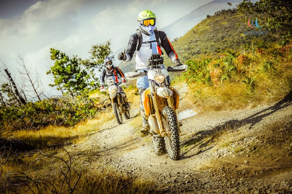 Two off-road motorcycles with riders wearing helmets and gear riding on a rocky dirt trail through a mountainous area with trees and bushes.